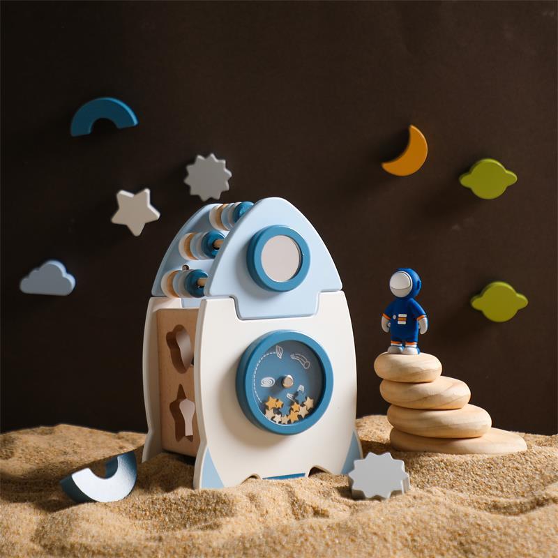 Wooden toy rocket with astronaut figure on a sandy surface with colorful shapes in the background
